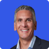 Smiling middle-aged man with gray hair wearing a dark blazer and light blue shirt against a blue background.